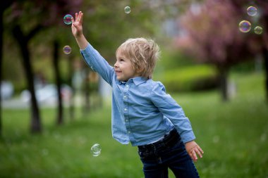 Sarışın bir çocuk, günlük kıyafetler içinde sevimli bir çocuk, parkta sabun köpükleriyle oynuyor, mutlu mesut koşuyor.
