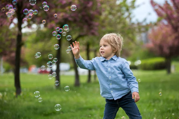 stock image Blond toddler child, cute boy in casual clothing, playing with soap bubbles in the park, running happily