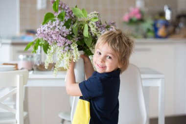 Güzel sarışın bir çocuğun yakın portresi, evde elinde leylaklarla vazo tutan küçük bir çocuk, evi dekore etmek için masanın üzerine koyuyor.