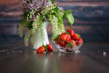 Esmer arka planda leylak ve çilek, çiçekli vazo ve meyveli kase.