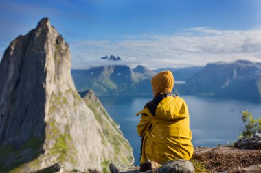 Sarı yağmurluklu bir kadın, bir kayanın üzerinde geriye doğru oturuyor ve Kuzey Norveç 'teki Senja Adası' ndaki Segla dağına bakıyor. Norveç 'in İskandinav topraklarında inanılmaz güzel manzara ve görkemli doğa