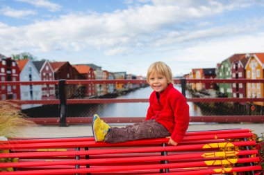 Tatlı sarışın çocuk, yaz boyunca Trondheim, Norveç 'i ziyaret ediyor, kırmızı bir bankta oturuyor, şehrin tadını çıkarıyor.