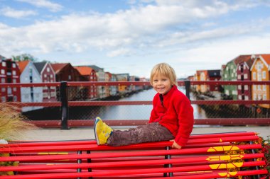 Tatlı sarışın çocuk, yaz boyunca Trondheim, Norveç 'i ziyaret ediyor, kırmızı bir bankta oturuyor, şehrin tadını çıkarıyor.