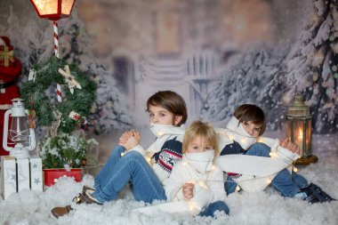 Çocuklar, karda oturuyorlar, tuvalet kağıdına sarılıyorlar ve Noel 'de hafif iplere sarılıyorlar, kameraya bakıyorlar.