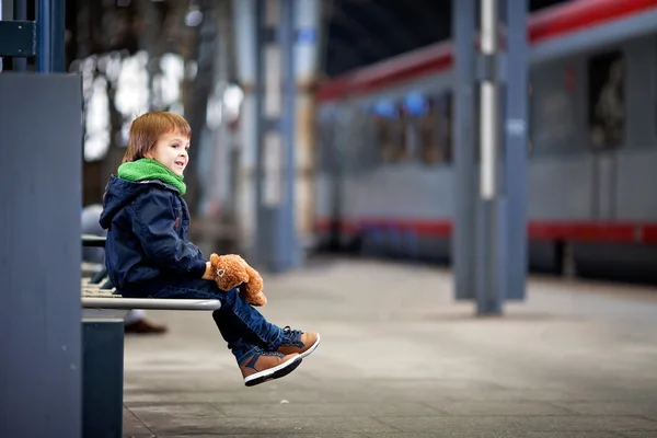 Lindo chico, sentado en un banco con oso de peluche, mirando un tren — Foto de Stock