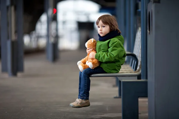 Leuke jongen, zittend op een bankje met teddy beer, kijken naar een trein — Stockfoto