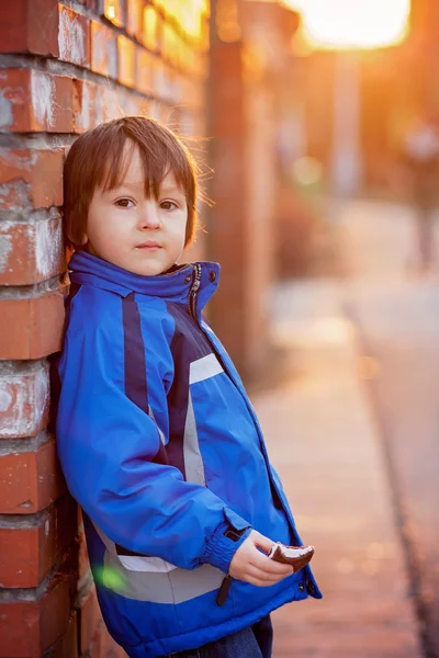 Adorable little boy, next to brick wall, eating chocolate bar on — Stock Photo, Image
