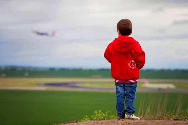 Cute boy in a red jacket, standing on a hill, watching airplane — Stock Photo, Image