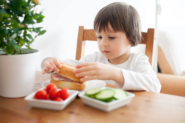 Vacker liten pojke, äta smörgås hemma, grönsaker på den — Stockfoto