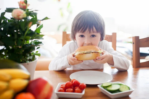 Vacker liten pojke, äta smörgås hemma, grönsaker på den — Stockfoto