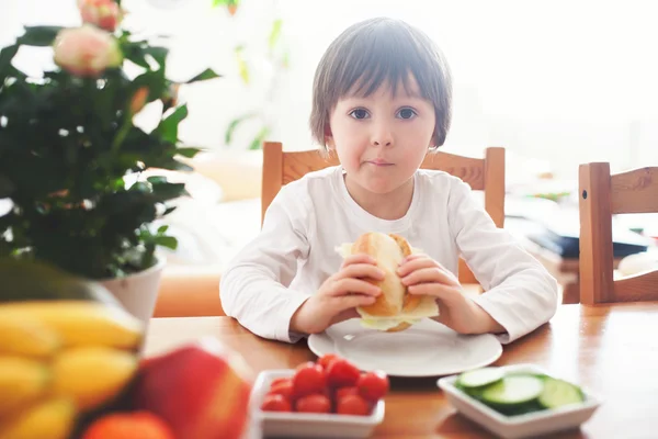 Vacker liten pojke, äta smörgås hemma, grönsaker på den — Stockfoto