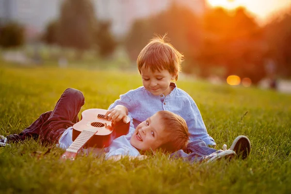 Два очаровательных мальчика, сидящие на траве, играющие на гитаре — стоковое фото