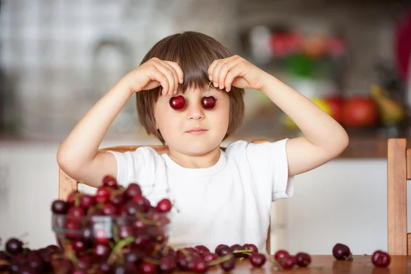 Cute little boy, eating cherries at home in the kitchen, making — Stock Photo, Image