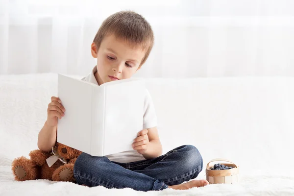 Süßer kleiner Junge, liest im Bett ein Buch und isst Blaubeeren, — Stockfoto
