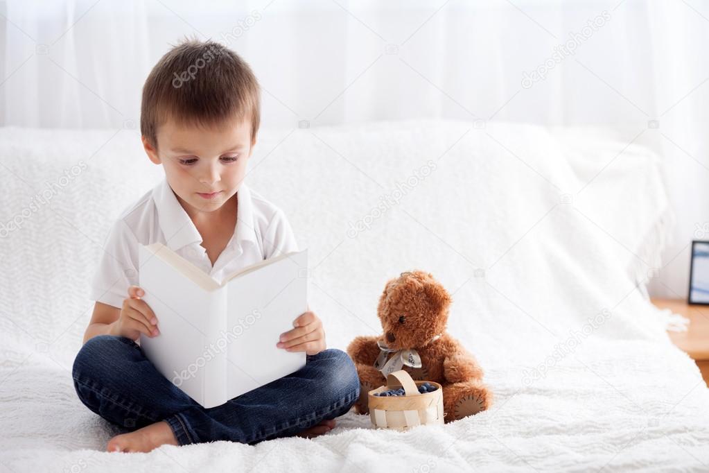 Sweet little boy, reading a book in bed and eating blueberries, 