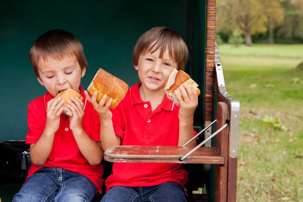 A két gyerek, egy védett padon ülve eszik szendvicseket — Stock Fotó