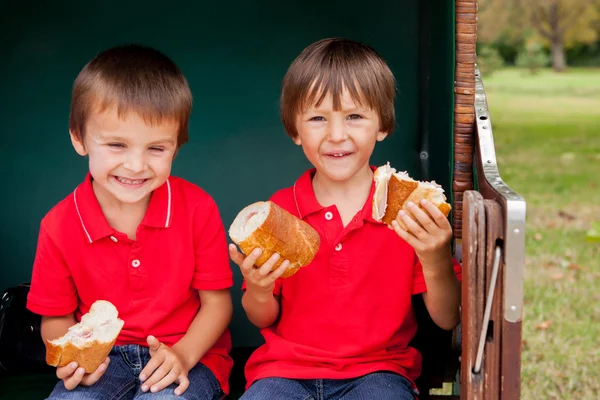 Duas crianças, sentadas em um banco protegido, comendo sanduíches — Fotografia de Stock