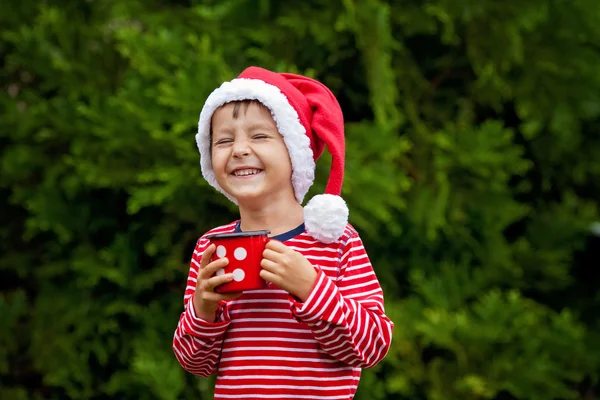 Doux garçon en chemise rayée avec chapeau de Père Noël, tenant tasse avec thé — Photo