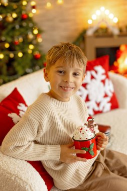 Sevimli anaokulu çocuğu, şöminenin önünde oturuyor, Noel hediyesini tutuyor ve evde kitap, eğlence ve mutluluk okuyor.