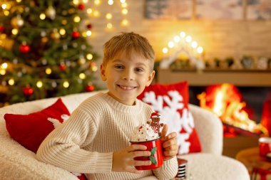 Sevimli anaokulu çocuğu, şöminenin önünde oturuyor, Noel hediyesini tutuyor ve evde kitap, eğlence ve mutluluk okuyor.