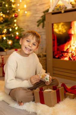 Sevimli anaokulu çocuğu, şöminenin önünde oturuyor, Noel hediyesini tutuyor ve evde kitap, eğlence ve mutluluk okuyor.