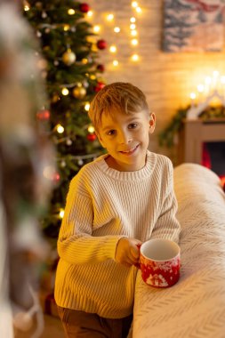 Sevimli anaokulu çocuğu, şöminenin önünde oturuyor, Noel hediyesini tutuyor ve evde kitap, eğlence ve mutluluk okuyor.
