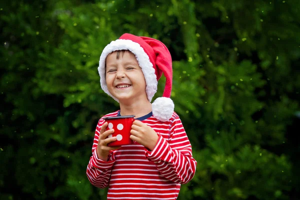 Menino doce em camisa listrada com chapéu de Papai Noel, segurando xícara com chá — Fotografia de Stock