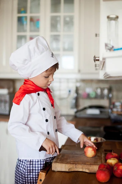 Sweet little boy, dressed as a chef, eating and cutting apples f — Stock Photo, Image