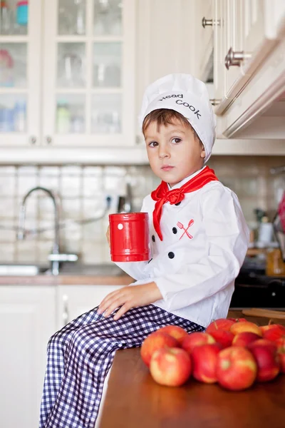 Sweet little boy, dressed as a chef, eating and cutting apples f — Stock Photo, Image