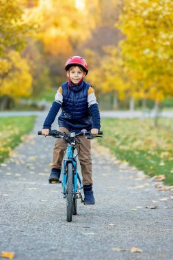 Sevimli küçük bir çocuk, renkli elbiseli bir çocuk, güneşli bir öğleden sonra bir sonbahar parkında bisiklet sürüyor, kameraya mutlu bir şekilde gülümsüyor.