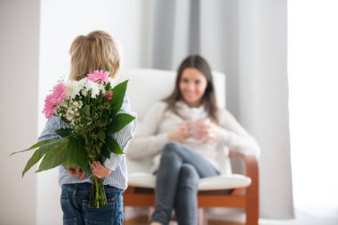 Güzel sarışın çocuk, güneşli oturma odasında anneler günü için küçük hediyeler vererek annesine çiçek ve kutu veriyor.