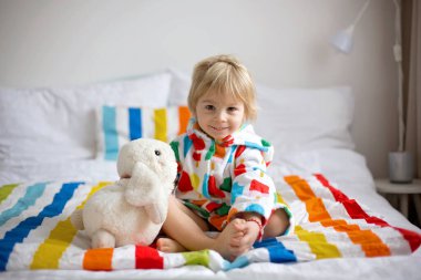 Mutlu çocuk, renkli bornozlu sarışın çocuk, içi doldurulmuş oyuncakla banyodan sonra yatakta oturuyor, gülümsüyor, renkli bir yatak.