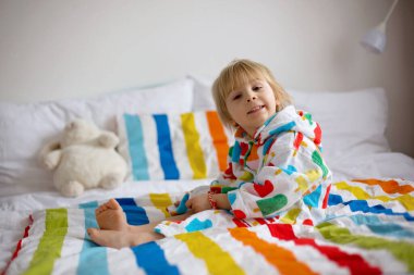 Mutlu çocuk, renkli bornozlu sarışın çocuk, içi doldurulmuş oyuncakla banyodan sonra yatakta oturuyor, gülümsüyor, renkli bir yatak.