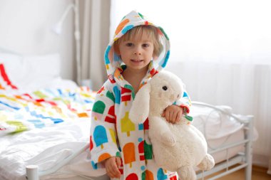 Mutlu çocuk, renkli bornozlu sarışın çocuk, içi doldurulmuş oyuncakla banyodan sonra yatakta oturuyor, gülümsüyor, renkli bir yatak.