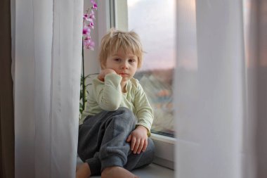 Sarışın çocuk, gün batımında pencerede oturuyor, dışarı bakıyor.