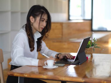 Kafedeki ahşap masada tabletle ödev yapan kız öğrencinin yan görüntüsü.