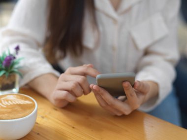 Kafede kahve fincanıyla rahatlarken akıllı telefon kullanan bir kadının görüntüsünü kapat.