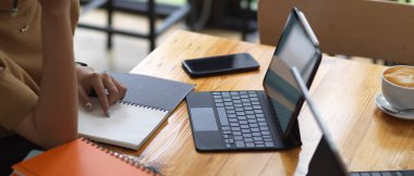 Tahta masa üzerinde tablet ve kırtasiyeyle ödev yapan kız öğrencinin kırpılmış görüntüsü.