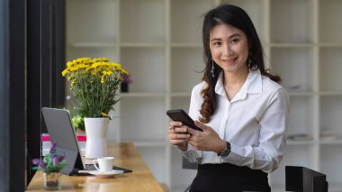 Ofisteki kameraya gülümserken akıllı telefonunu tutan kadın ofis çalışanı. 