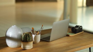 Ofis odasındaki ahşap masada dizüstü bilgisayar, kamera ve dekorasyonla kesilmiş çalışma alanı görüntüsü.