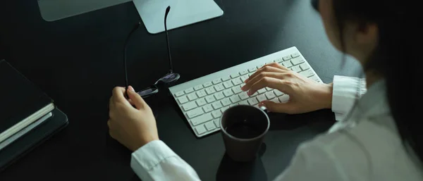 Overhead shot of female hand holding eyeglasses and typing on computer keyboard on office desk
