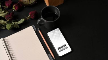 Akıllı telefon, defter, kahve fincanı ve çiçekli çalışma alanının en üst görüntüsü.