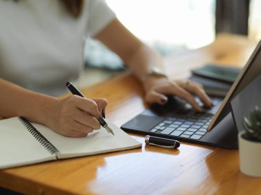 Ofis odasındaki ahşap masada tablet ve kırtasiyeyle çalışan kadın elinin yakın görüntüsü.