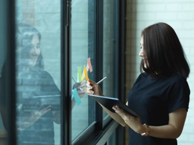 Ofisteki cam duvarda dijital tablet ve yapışkan notla çalışan kadın ofis çalışanının yan görüntüsü