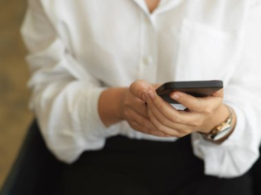 Ofis odasında otururken akıllı telefon kullanan bir kadın elinin kırpılmış görüntüsü.