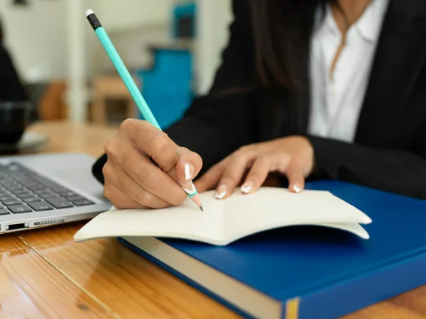 Close up view of female hand with pencil writing on notebook while working with laptop on wooden table