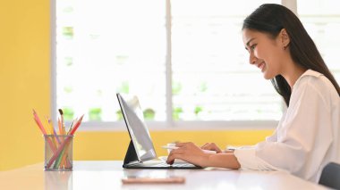 Kütüphanedeki masada dijital tabletle ödev yapan genç kız öğrencinin yan görüntüsü