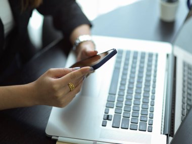 Ofisteki masada dizüstü bilgisayarla çalışırken akıllı telefon kullanan bir iş kadını görüntüsü.