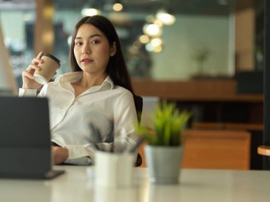Ofiste arka koltukta rahatça uzanırken elinde kağıt bardakla kameraya bakan kadın ofis çalışanının portresi.
