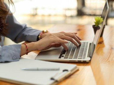 Tahta ofis masasındaki dizüstü bilgisayarda kağıt ve malzemelerle yazı yazan kadın elleri yan görünüm 